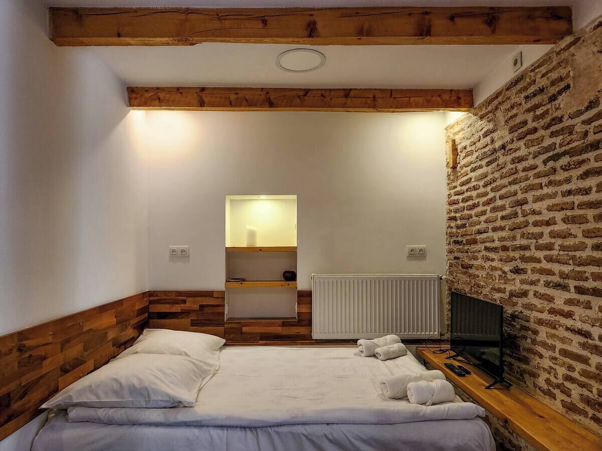 A bedroom with a wooden headboard, a bed with white linens, a bedside table with a lamp, a brick wall, and a radiator.