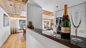 A modern kitchen with a wooden ceiling, a bar area with a bottle of Moët & Chandon champagne and two glasses, and a view of the living area with a dining table and chairs. - Chalet Larchwood by Mrs Miggins 3 (Grimentz)