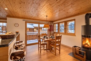 A cozy room with a wooden ceiling, a dining table with chairs, a fireplace, and a view of a snowy landscape through the windows.