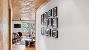 Un pasillo con suelo de madera y una pared blanca decorada con cuadros enmarcados. Una puerta de madera da a una habitación con televisor y vistas a árboles a través de una ventana.