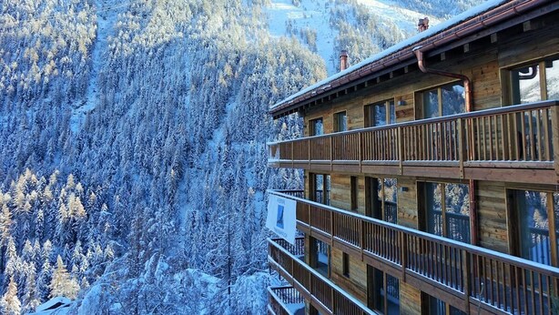 A wooden building with multiple balconies overlooking a snow-covered forest and mountains.