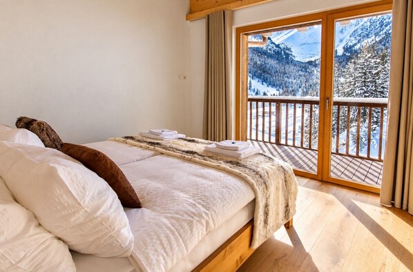 A bedroom with a bed, pillows, a blanket, and a view of a snowy mountain landscape through a large window.