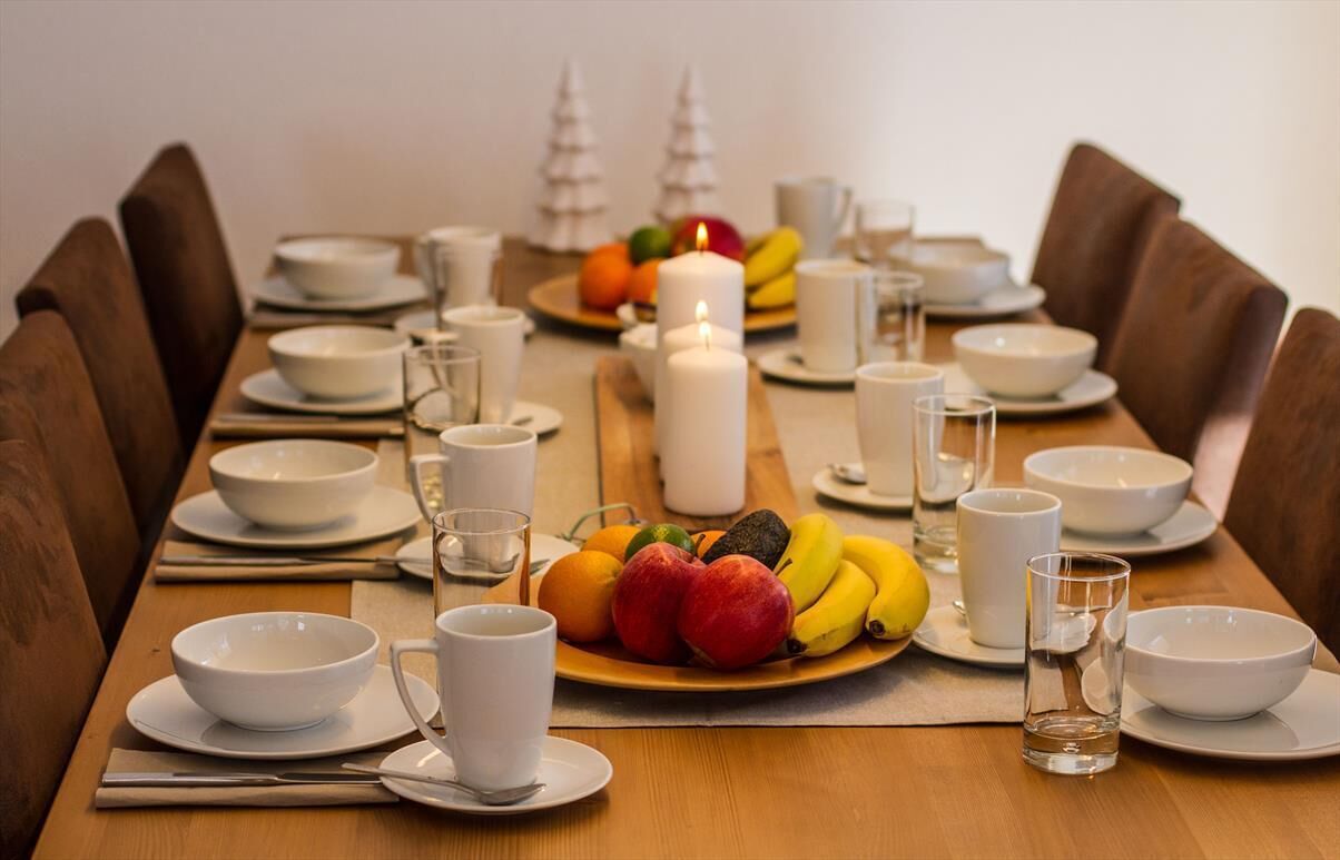 Mesa de comedor decorada con cuencos, platos y cubiertos blancos, acompañada de frutas y una vela.
