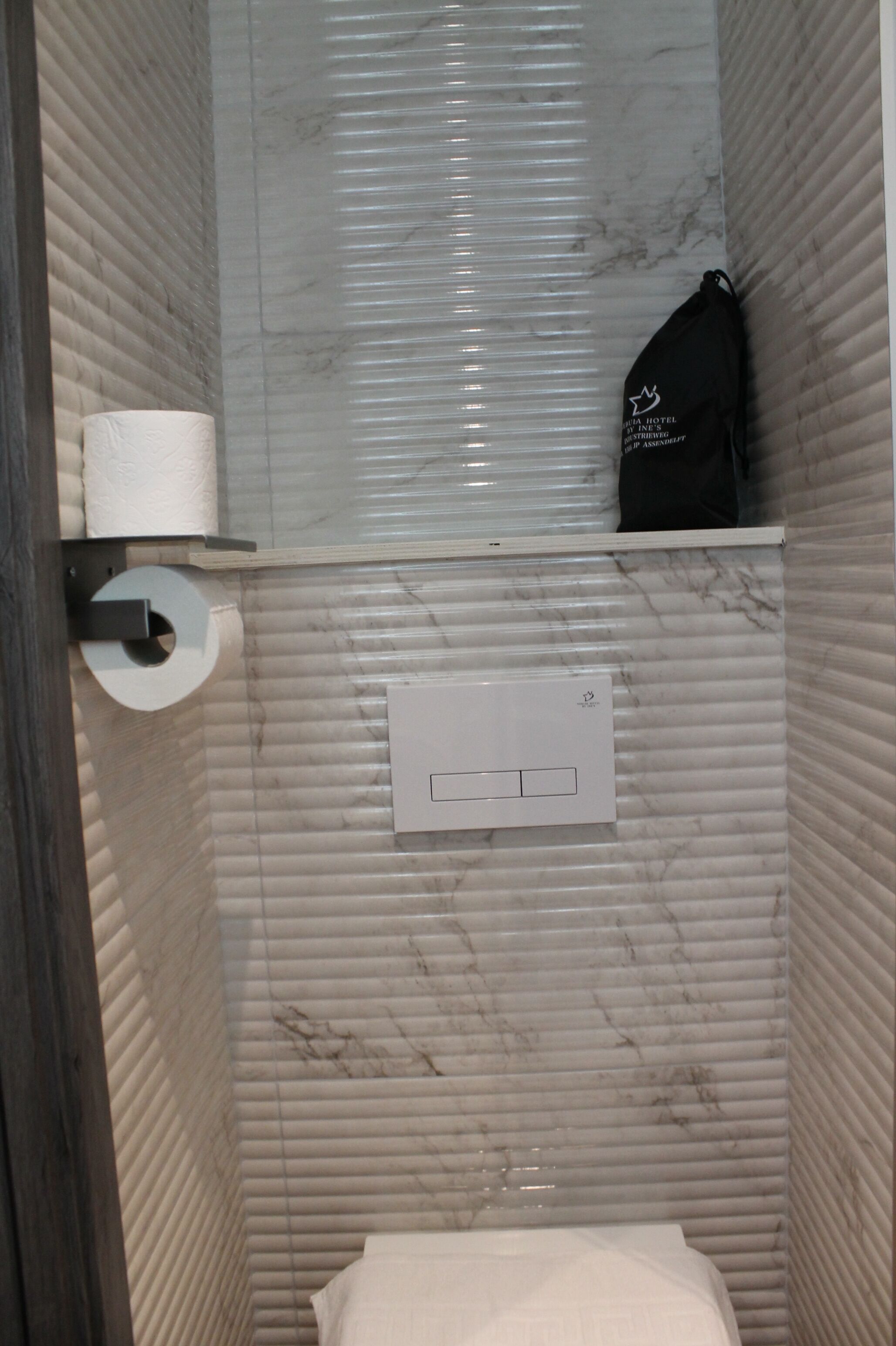 A bathroom with a toilet, a paper towel holder, a black bag, and a light switch on the wall.