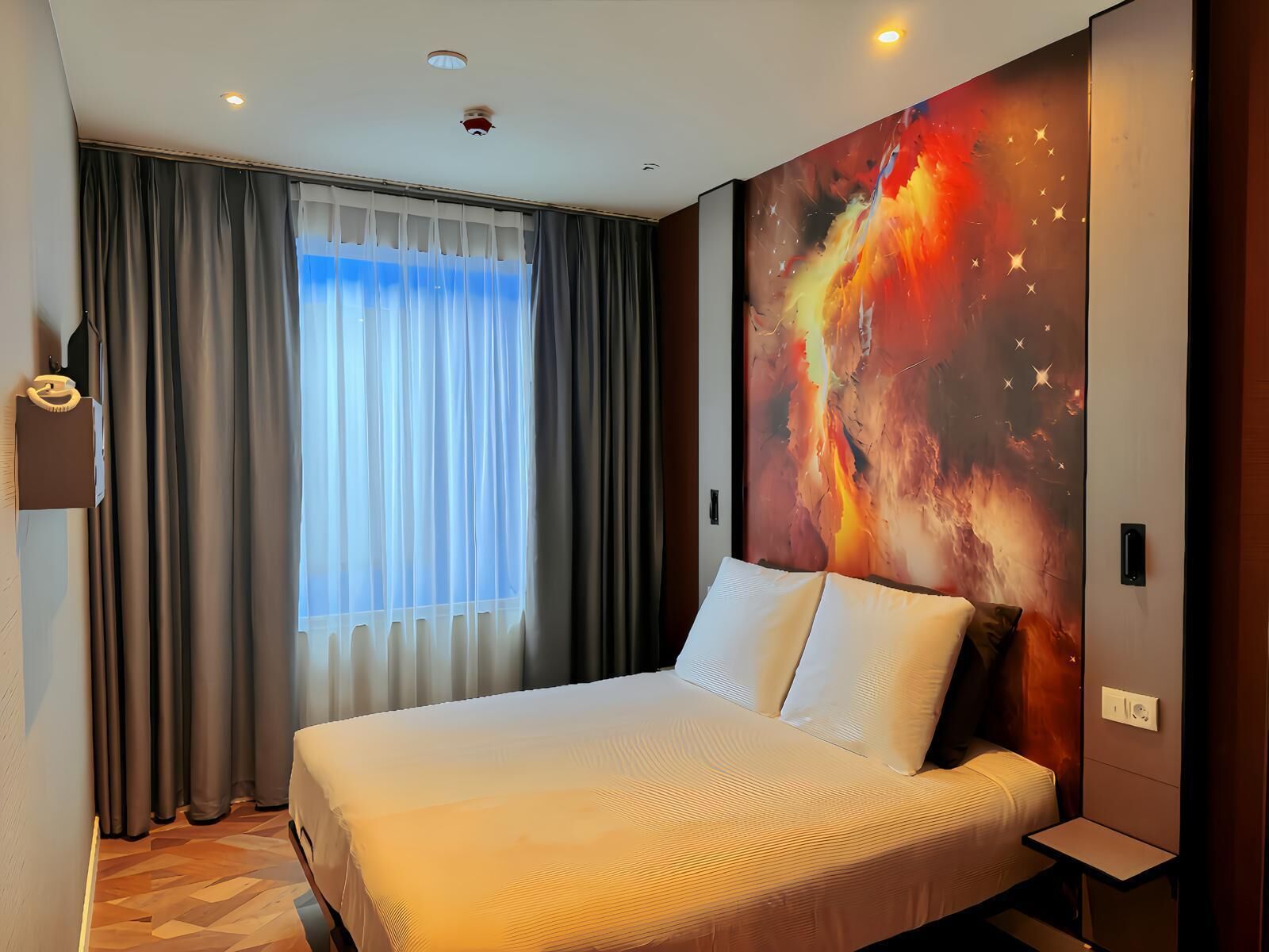 A hotel room with a large bed, a view of the sky through sheer curtains, and a vibrant abstract wall mural.