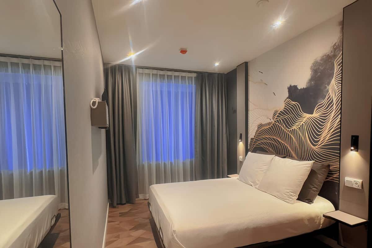 A modern hotel room with a large bed, a mirror, a wall-mounted artwork, and a window with curtains.
