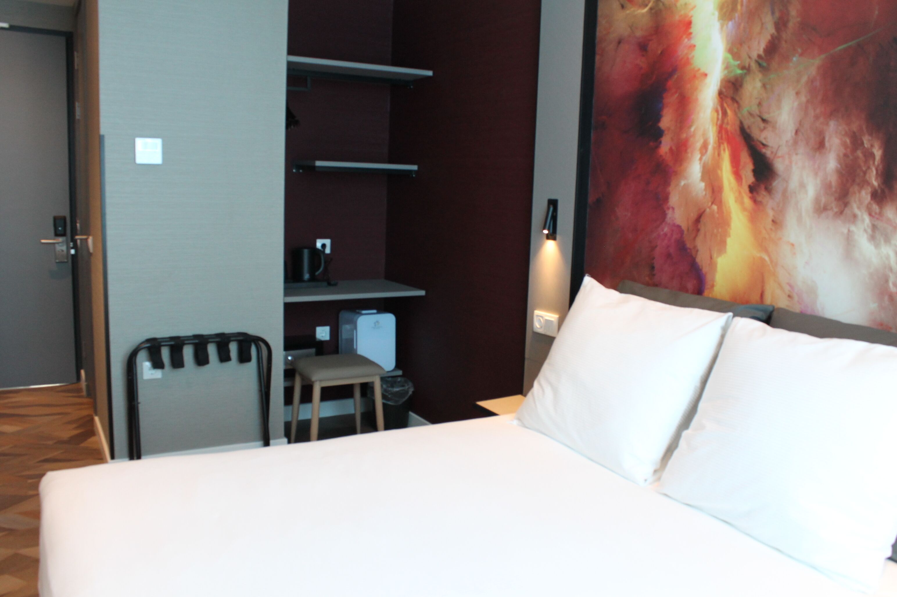 Habitación de hotel con cama, escritorio y una silla. Se observa una estantería con hervidor y un cubo de basura.