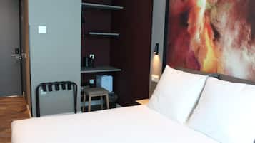Habitación de hotel con cama, escritorio y una silla. Se observa una estantería con hervidor y un cubo de basura.