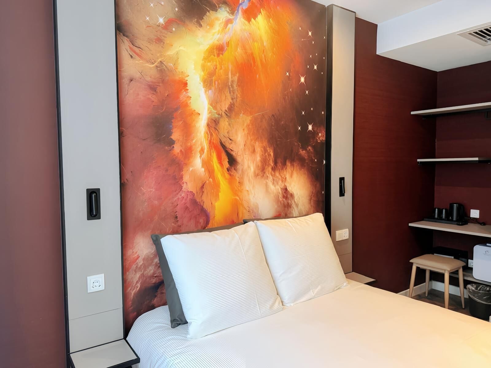 A modern hotel room with a large abstract painting on the wall, a bed with white linens, a small wooden stool, and a desk with a computer.