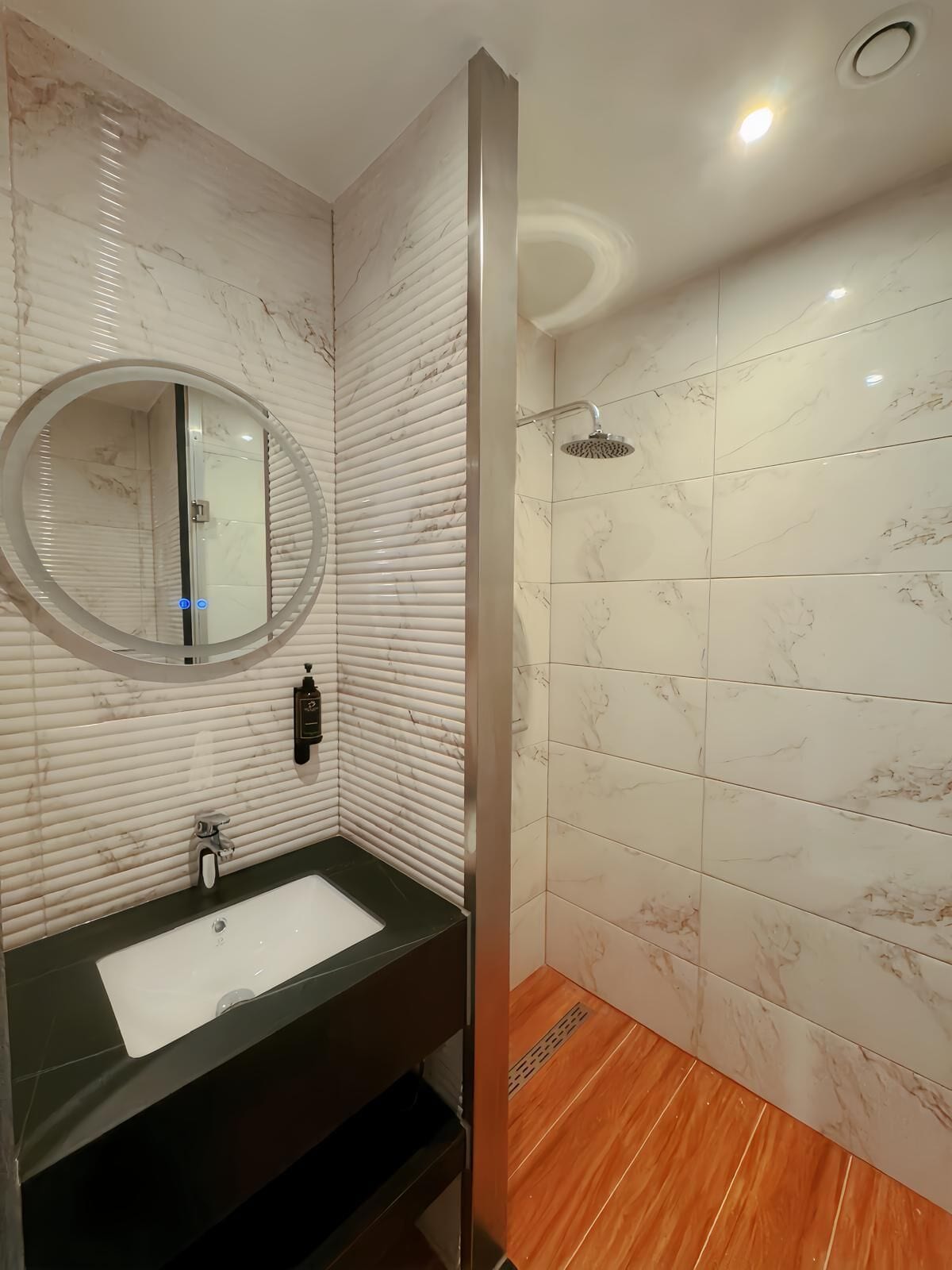 A bathroom with a marble vanity, a round mirror, a shower with a handheld showerhead, and a wooden floor.