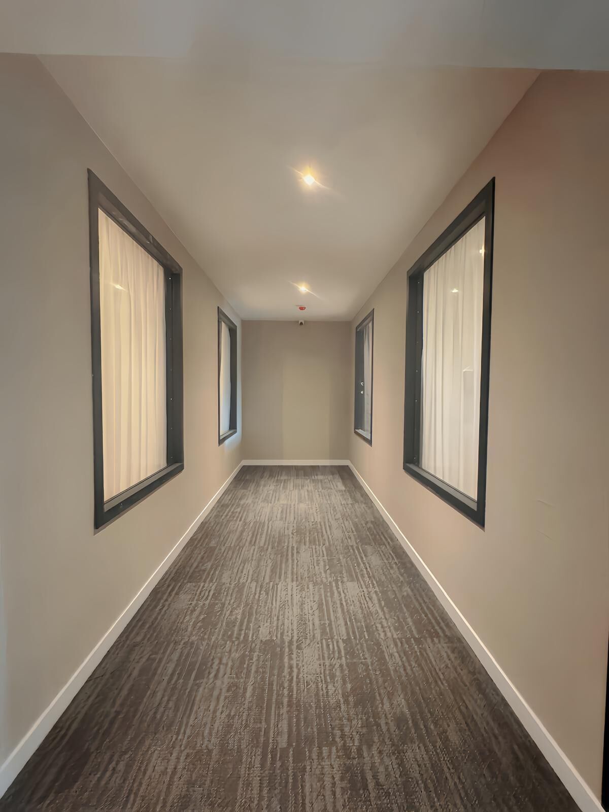 Un couloir aux murs de couleur beige, de grandes fenêtres ornées de rideaux blancs et un plancher en bois.