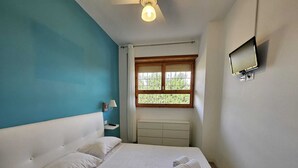 Een slaapkamer met een bed, nachtkastje, lamp, televisie en een raam met uitzicht op groen.