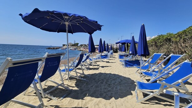 Een strand met rijen blauwe ligbedden en bijpassende parasols, een heldere lucht en de zee op de achtergrond.