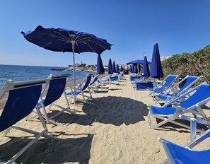 Een strand met rijen blauwe ligbedden en bijpassende parasols, een heldere lucht en de zee op de achtergrond.