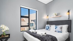 Ein modernes Schlafzimmer mit großem Fenster, einem Bett mit gerollten Handtüchern, einem kleinen Tisch mit Pflanze und Wandleuchten.