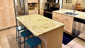 Uma cozinha com armários de madeira, eletrodomésticos de aço inoxidável, bancada de granito e banquetas azuis.