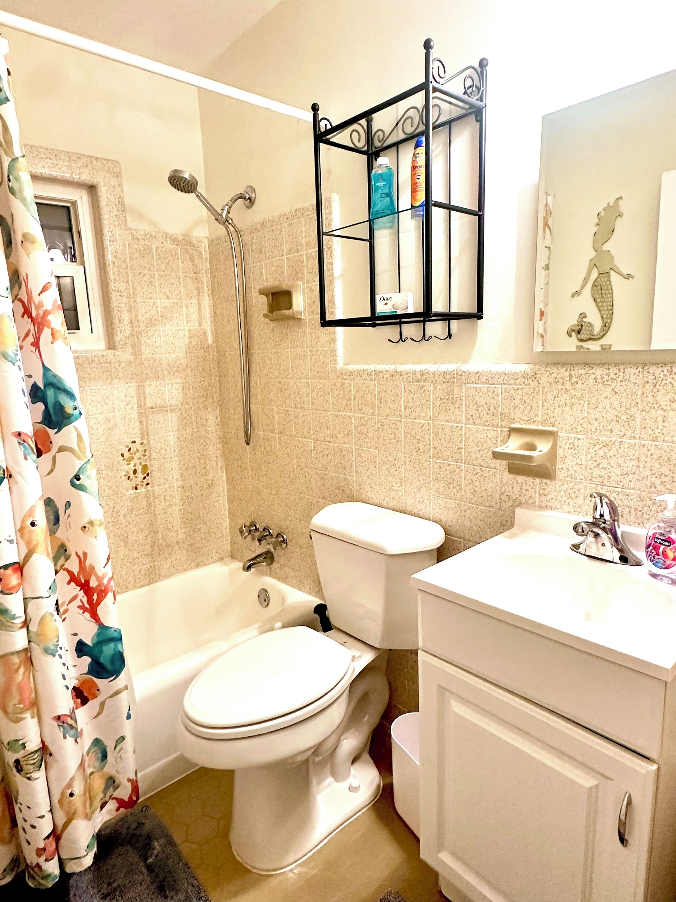 Un baño con bañera, inodoro y lavamanos. Cuenta con un cabezal de ducha, un perchero y un mural decorativo con una sirena.