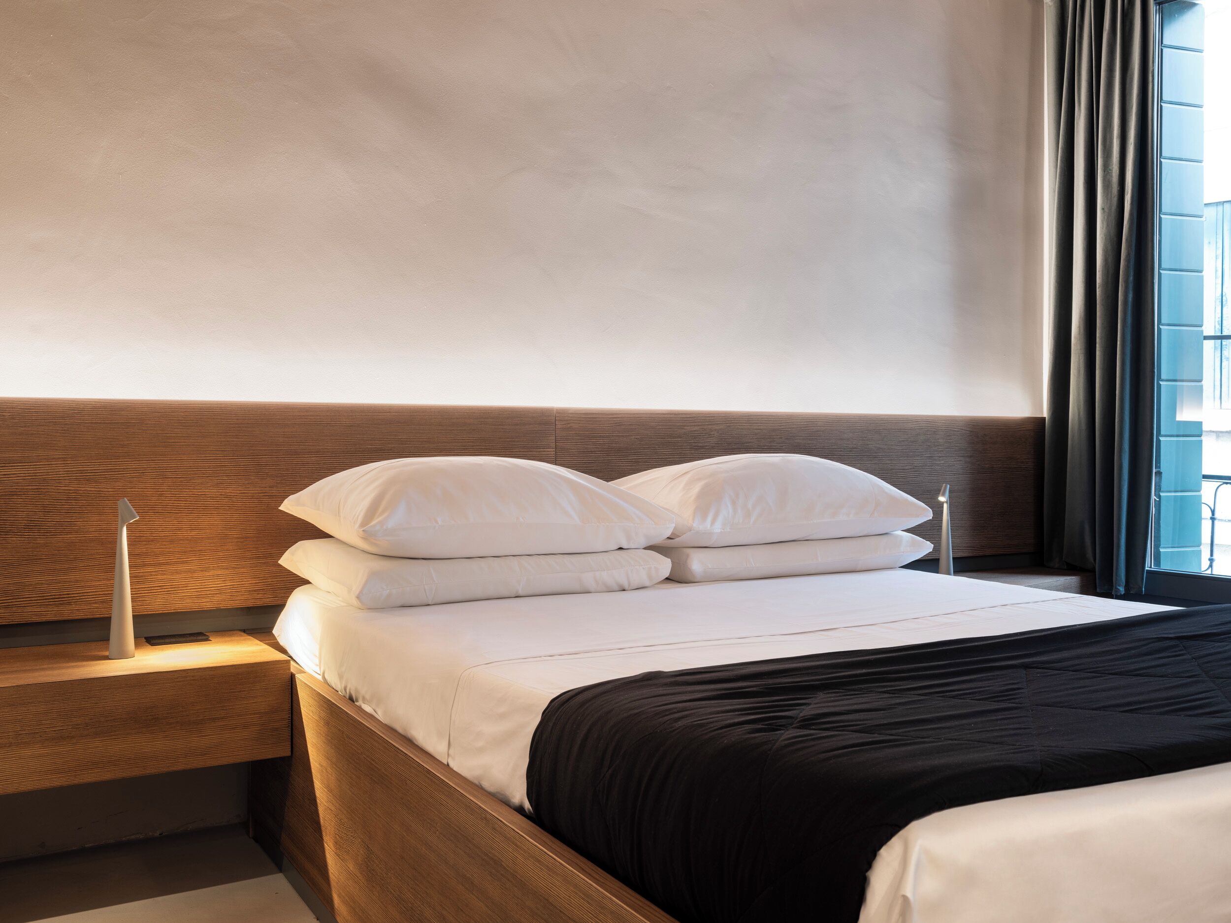Una cama bien tendida con ropa blanca y un plaid negro, un cabecero de madera y una mesita de noche con una lámpara.