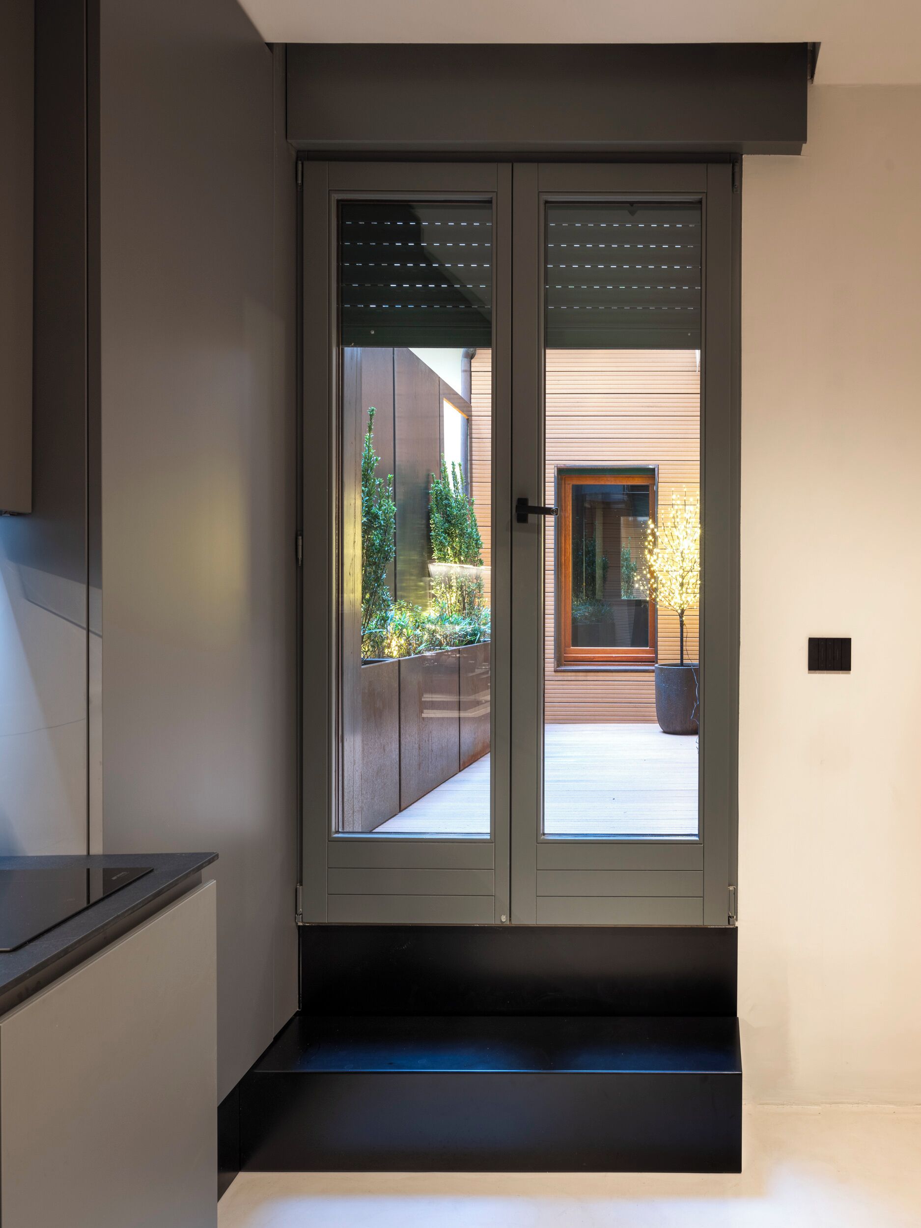 Un ingresso moderno con una porta in vetro, una piccola area esterna con una pianta in vaso e una panchina in cemento.