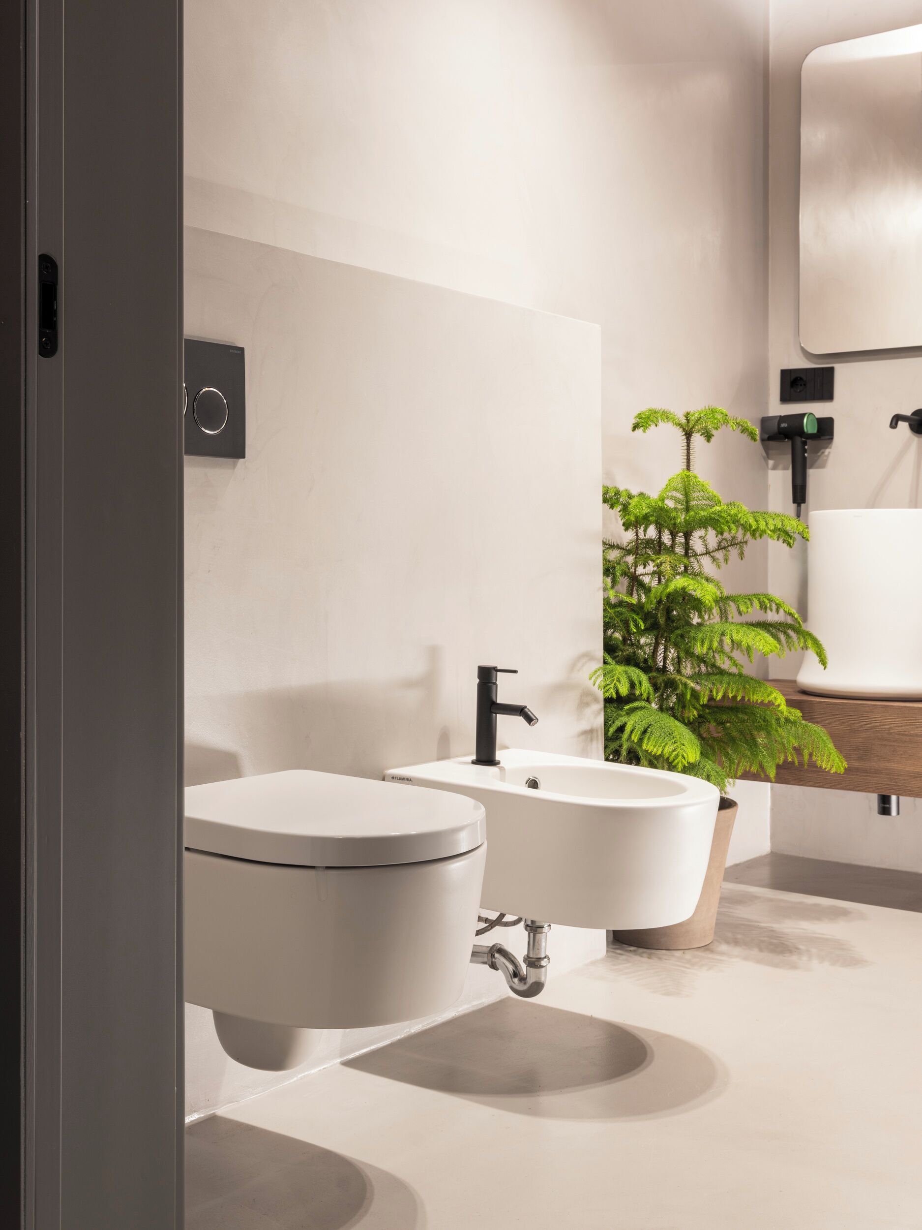 Un baño moderno con inodoro, lavamanos y bidé. Hay una planta y un espejo en la pared.