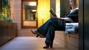 Una mujer sentada en un banco leyendo un libro en un espacio interior moderno con suelo de madera y una luz cálida de fondo.