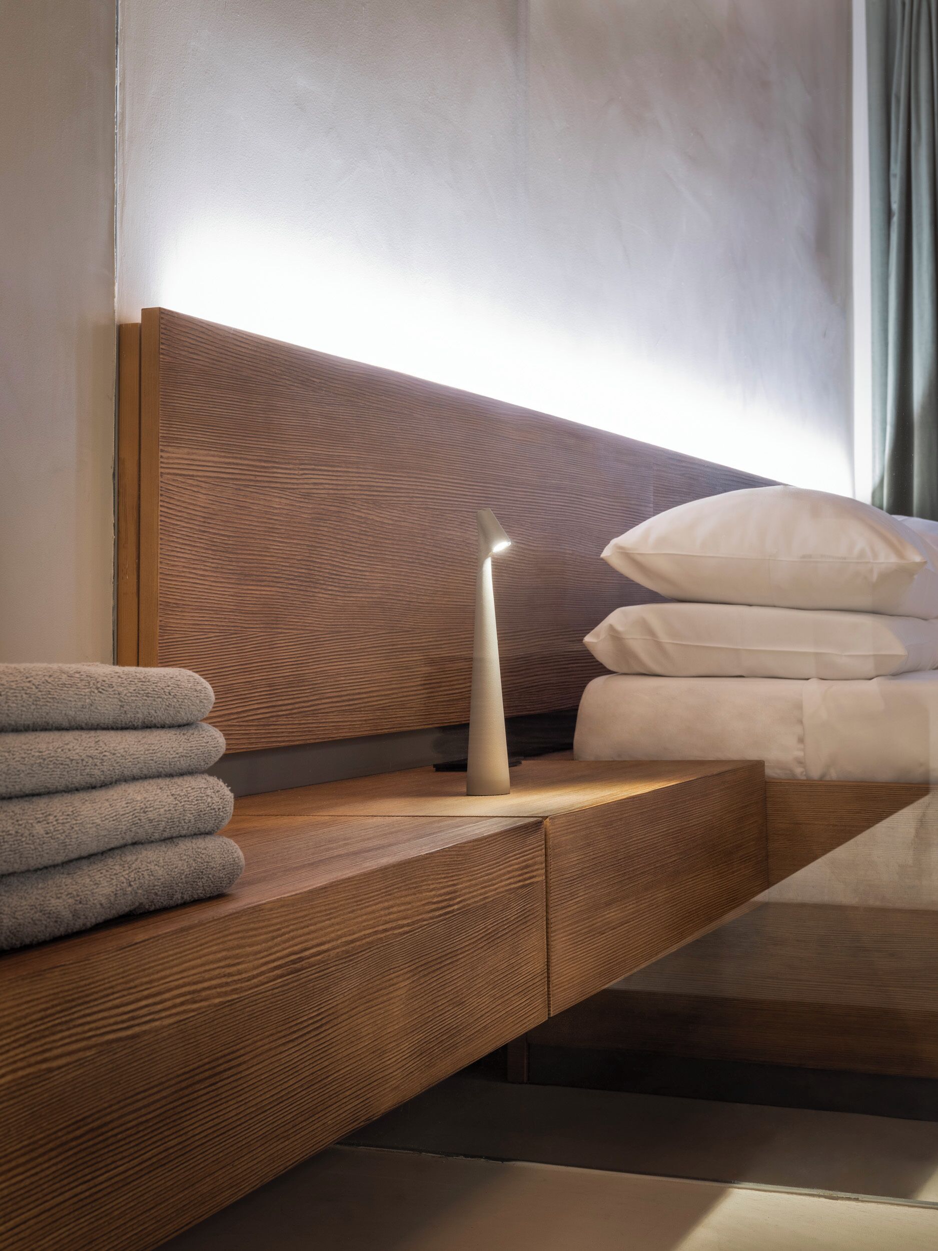 A modern bedroom with a wooden headboard, a bedside table with towels, and a bedside lamp.