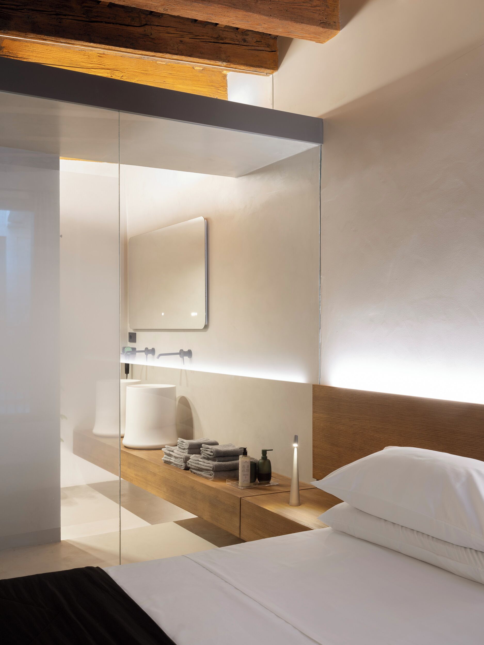 A modern hotel room with a wooden headboard, a bedside table with toiletries, and a large mirror.