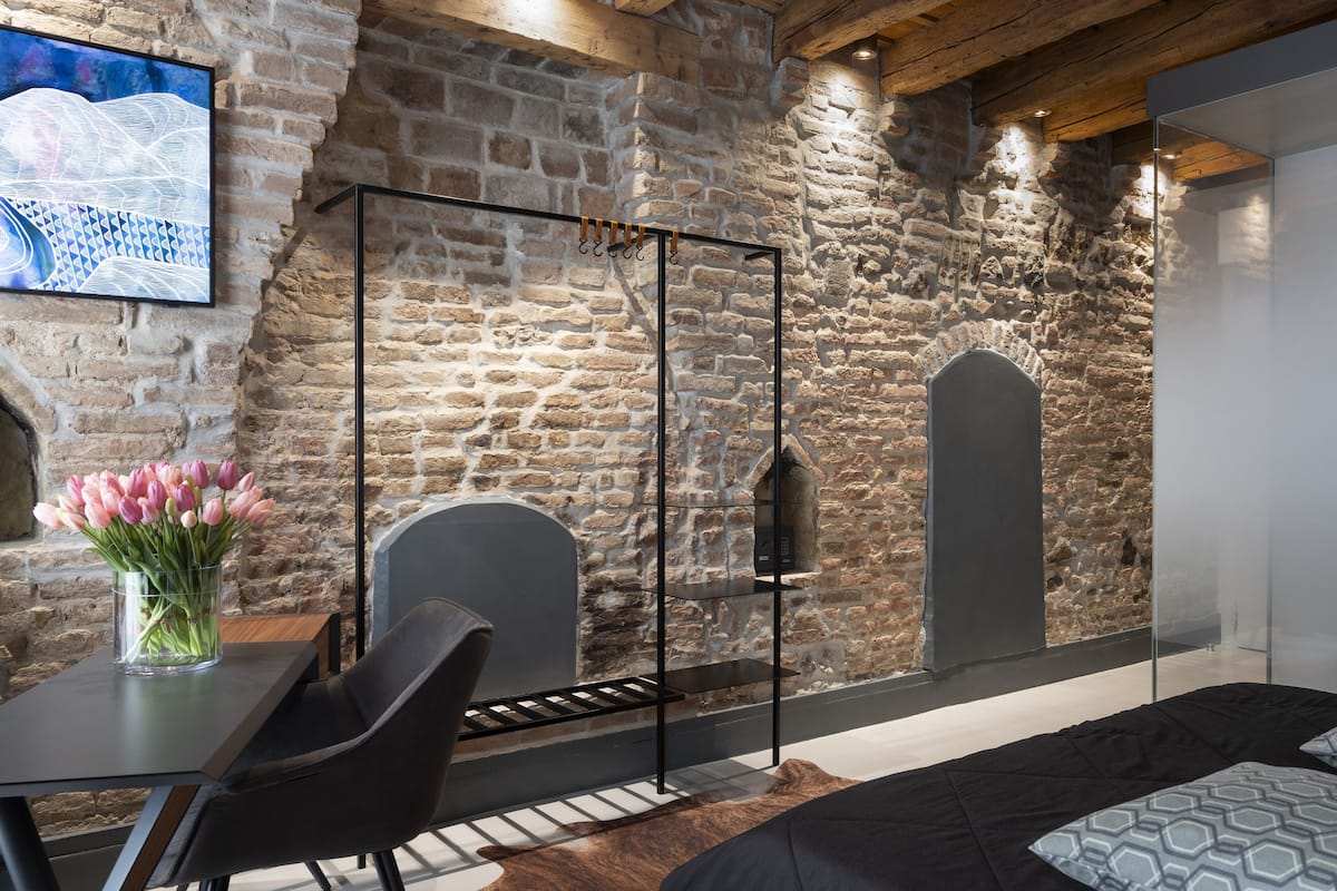 A modern room with exposed stone walls, a bed, a desk with a chair, and a vase of flowers.