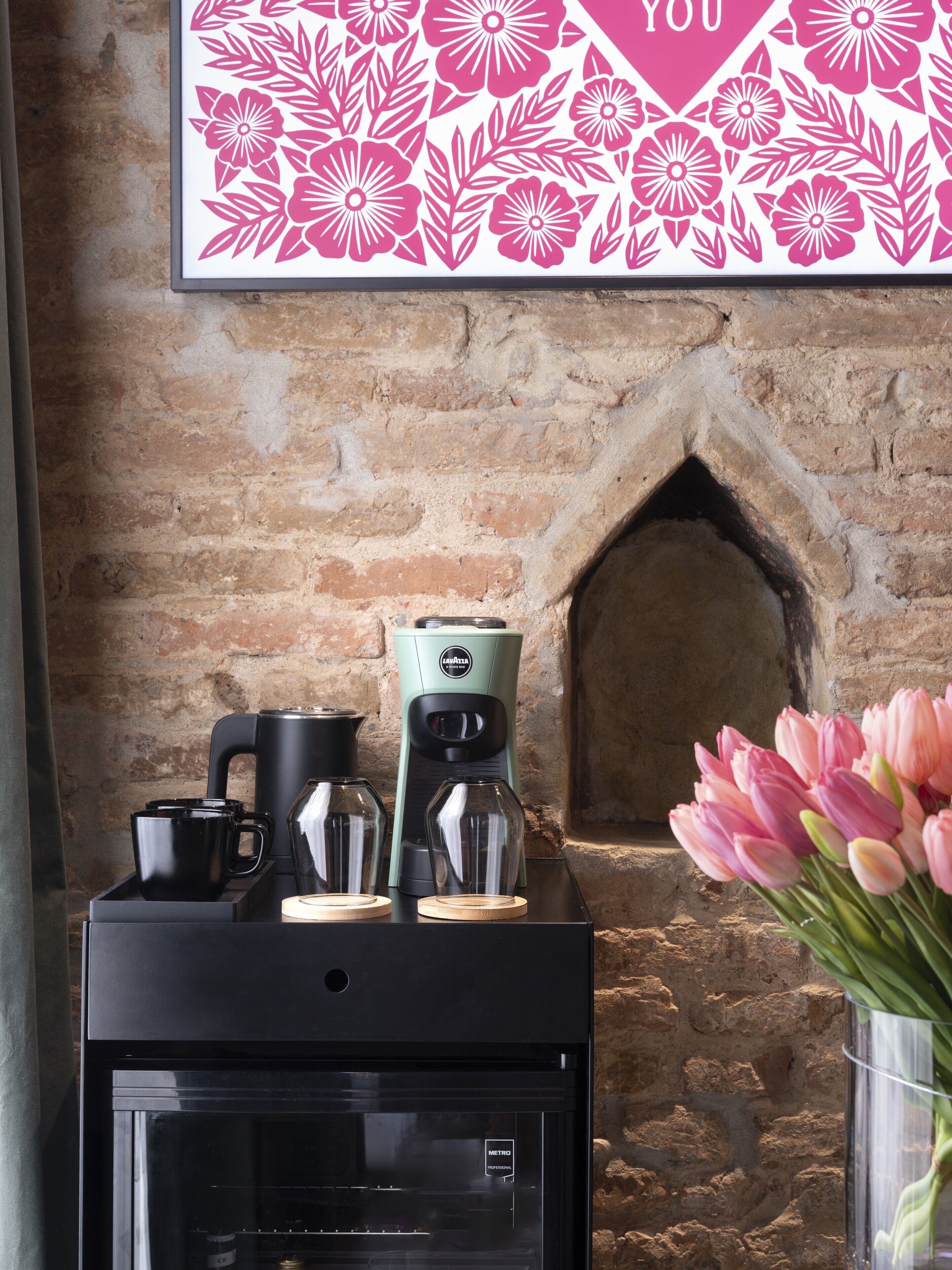 A coffee machine with a glass carafe and a coffee pot on a tray, a glass of water, and a vase with pink tulips against a brick wall.