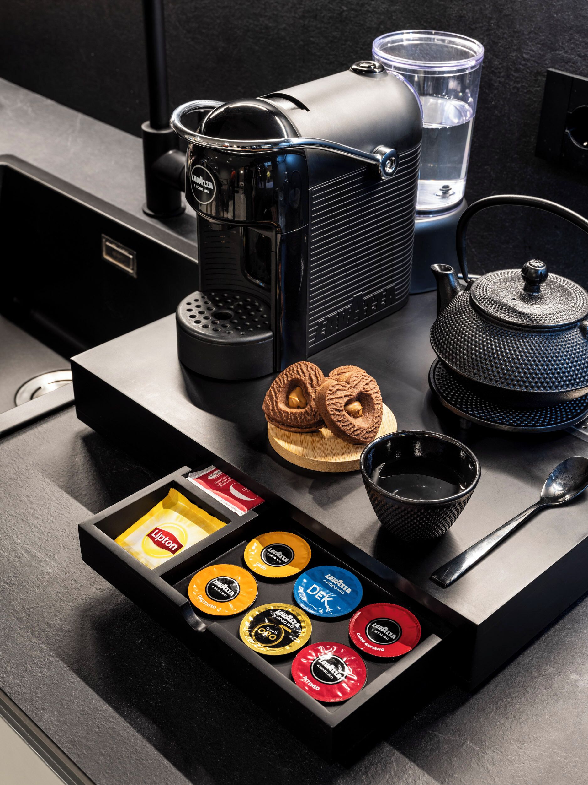 A coffee machine, a teapot, a cup, a spoon, and a drawer with coffee capsules.