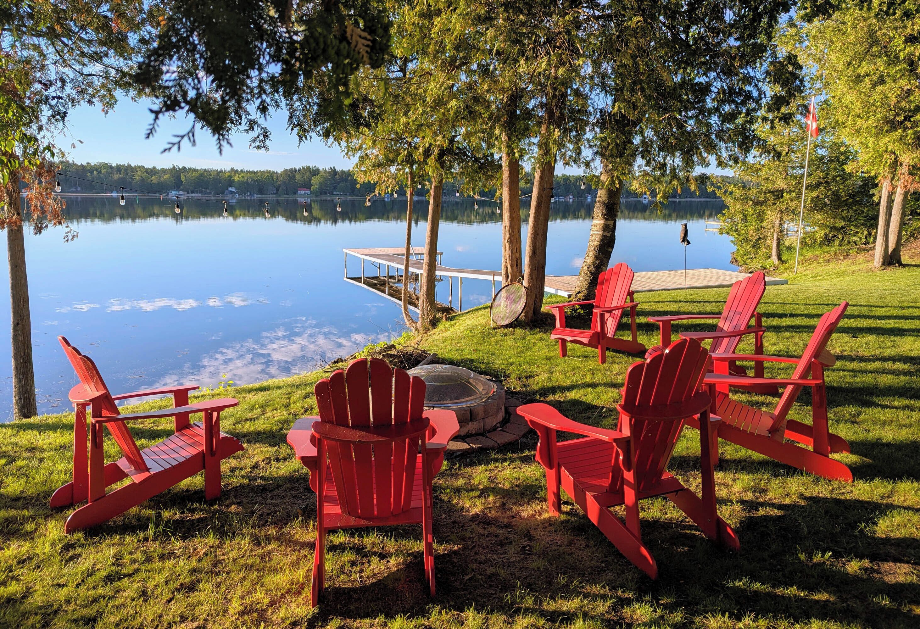 A lakeside setting with red Adirondack chairs, a fire pit, and a dock extending into the water.