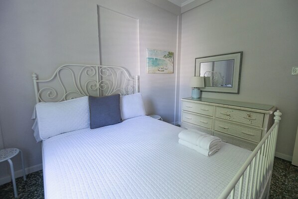 A neatly made bed with a white headboard and pillows, a bedside table with a lamp, and a dresser with drawers.