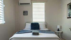 Un lit bien fait, avec un oreiller bleu, une serviette pliée et une couverture bleue. On y trouve une petite table de chevet avec une lampe, et un tableau encadré est accroché au mur.