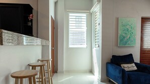 Un couloir avec un canapé bleu, des tabourets en bois, une porte et une fenêtre avec des stores.