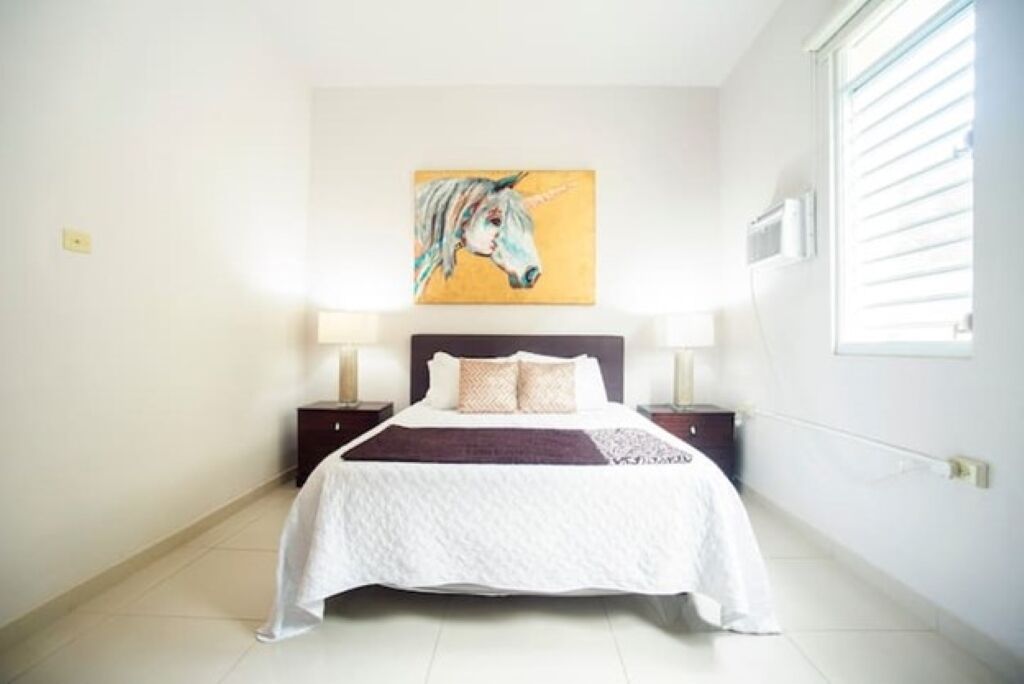 A bedroom with a bed, two bedside tables, a painting on the wall, and a window with blinds.