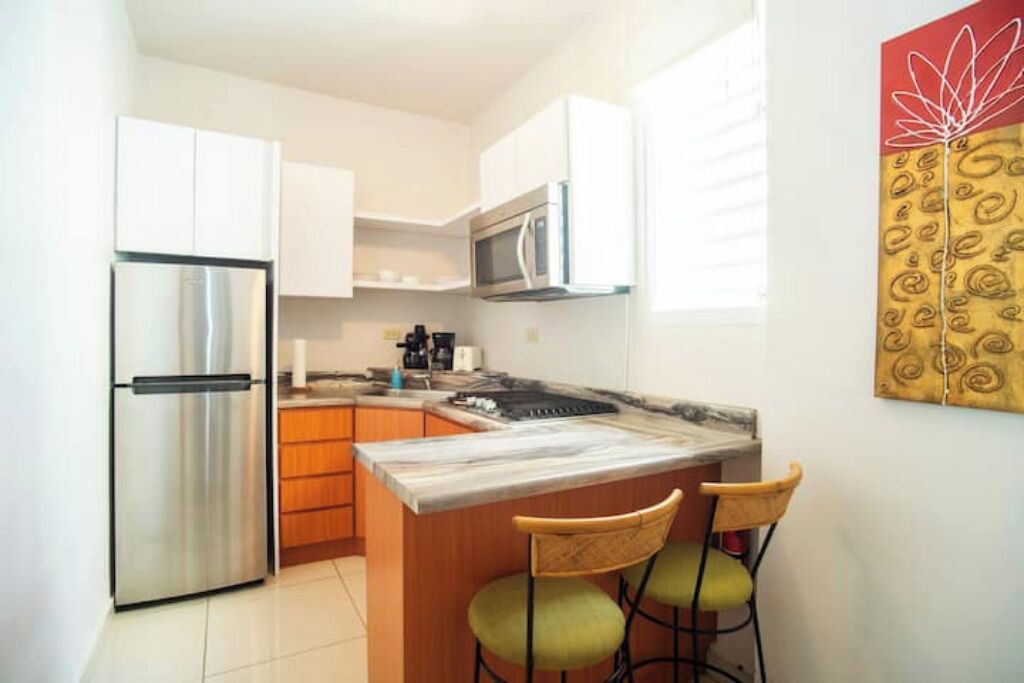 A compact kitchen with white cabinets, a stainless steel refrigerator, a microwave, a coffee maker, and a marble countertop. There are two green chairs at the island.