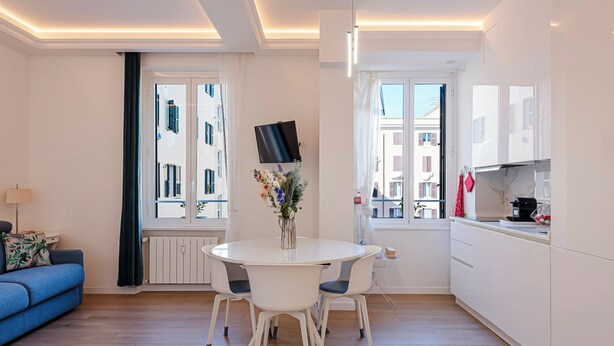 Una cucina moderna con mobili bianchi, un tavolo da pranzo con sedie, un televisore sopra il tavolo e una finestra con vista sugli edifici.