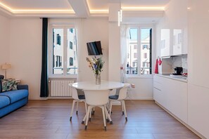 A modern kitchen with white cabinets, a dining table with chairs, a TV mounted above the table, and a window with a view of buildings.