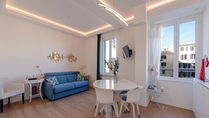 Un soggiorno moderno con un divano blu, sedie bianche, un tavolo rotondo e un'ampia finestra che si affaccia su edifici.