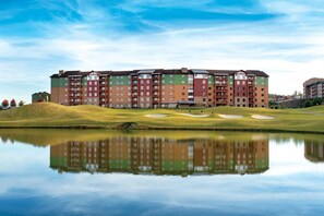 Un hôtel à plusieurs étages dont l’image se reflète dans une étendue d’eau calme, entouré d’une pelouse bien entretenue et de bunkers de sable.