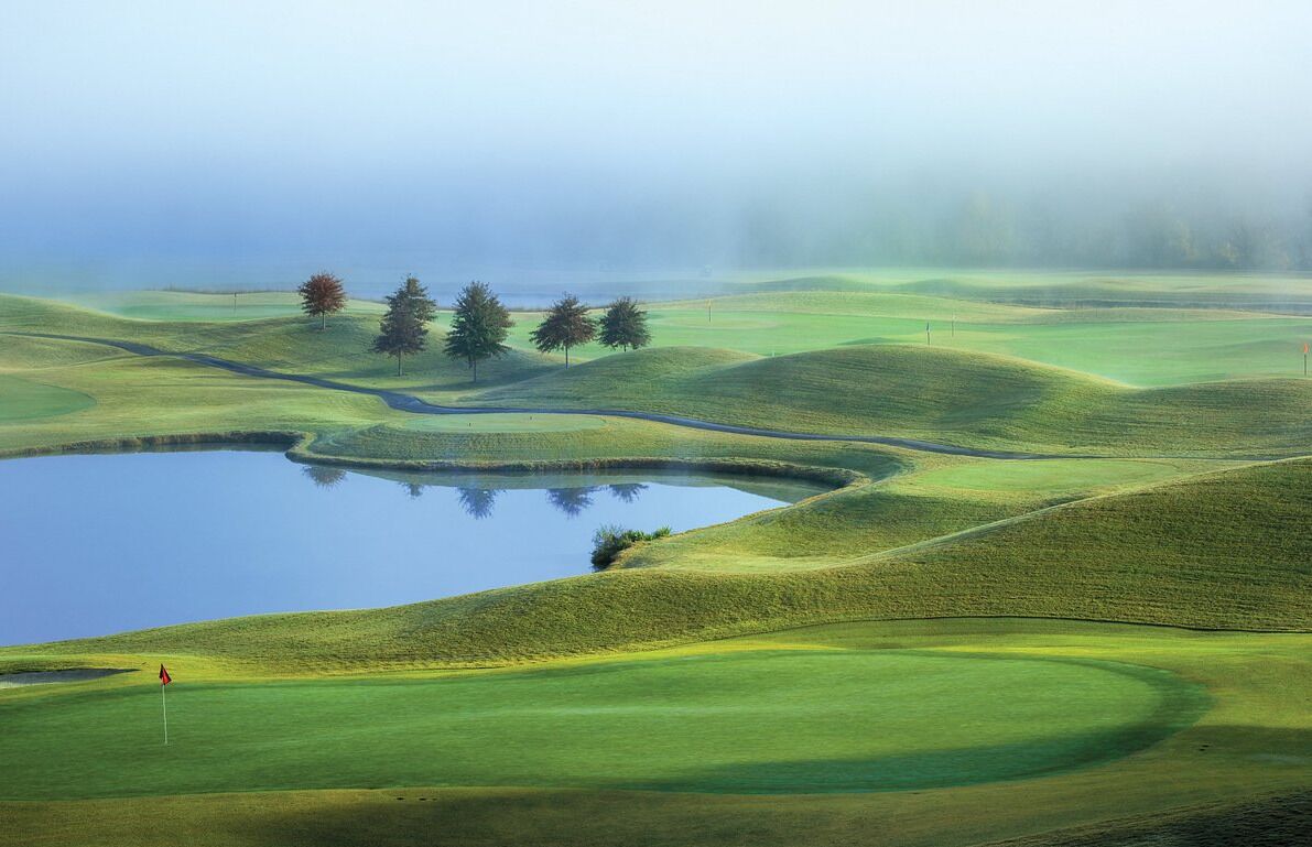 A golf course with rolling green hills, a pond, and a few trees.