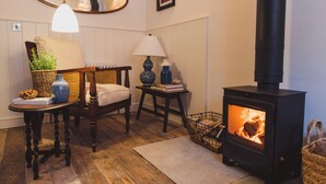 A cozy room with a wooden floor, a chair, a table with a lamp, a bookshelf, and a wood-burning stove. - Pebble Cottage 3, Buckingham Mews (Broadstairs)