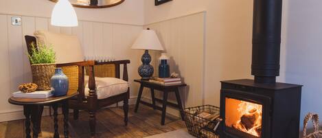 A cozy room with a wooden floor, a chair, a table with a lamp, a bookshelf, and a wood-burning stove.