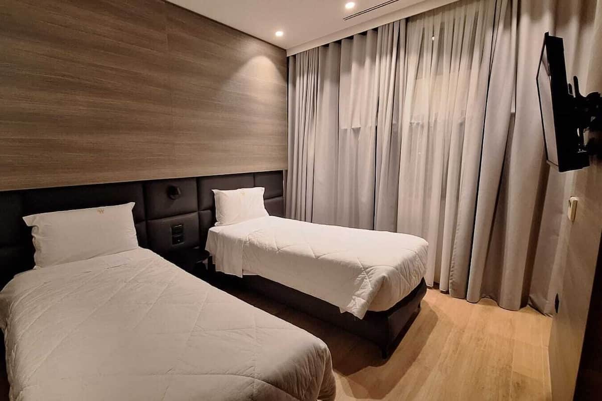 A hotel room with two beds, a dark headboard, a flat-screen TV mounted on the wall, and a large window with curtains.