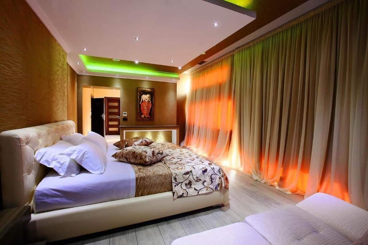 A modern bedroom with a large bed, a bedside table, and a headboard. There is a door leading to another room and a painting on the wall.