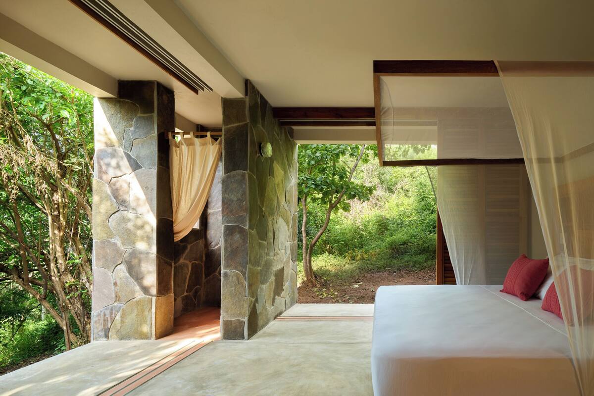 Une chambre avec un mur en pierre, un lit avec une literie blanche et rouge, et une vue sur la verdure à l ’extérieur.