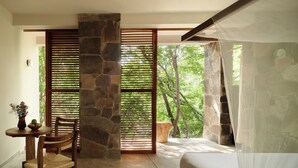 A bedroom with a stone wall, a bed with a canopy, a wooden chair, a small table with a vase and fruit, and a view of trees through a window with wooden blinds. - Cocolia Hotel (Santa María Tonameca)