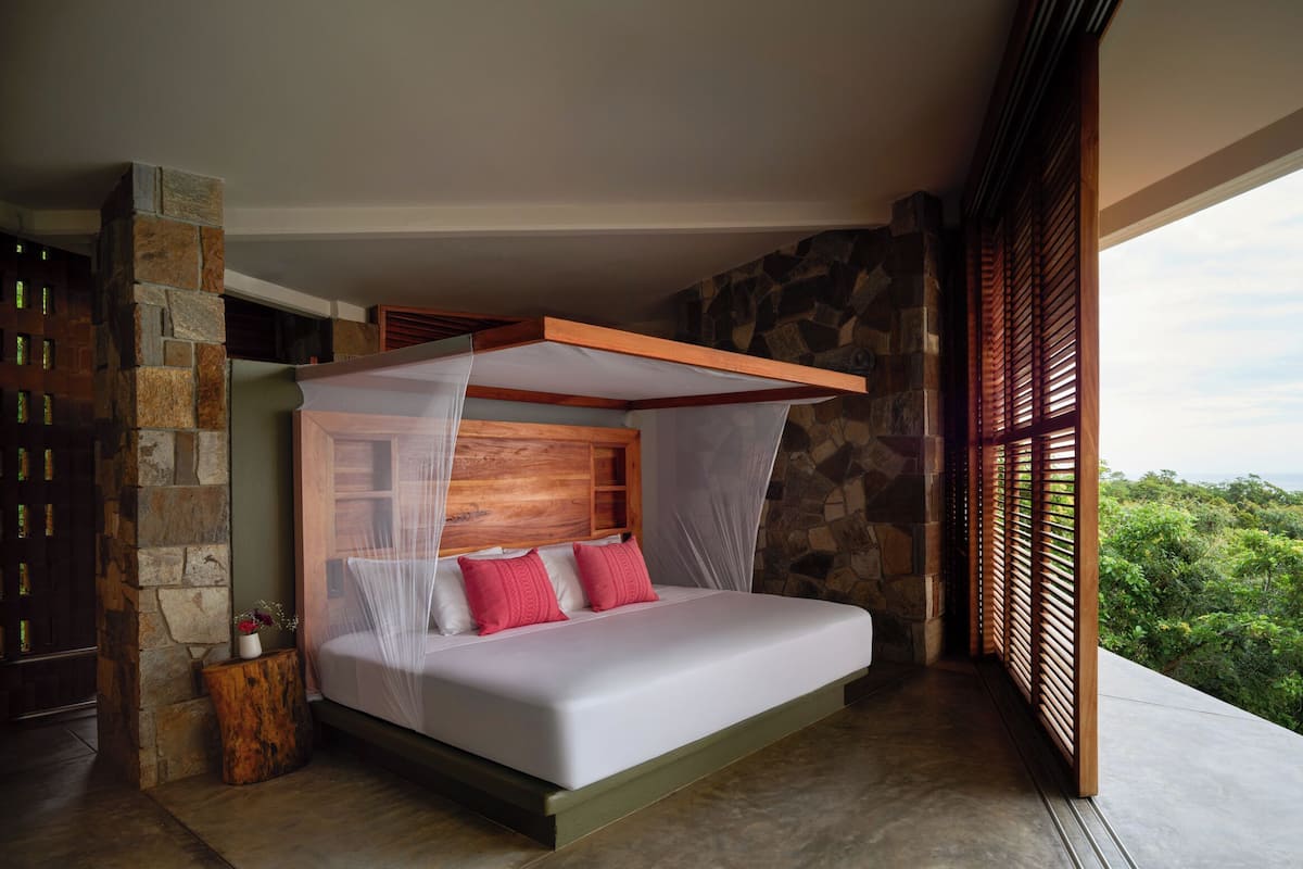 Une chambre avec un mur en pierre, une tête de lit en bois, un lit avec une literie blanche et des oreillers rouges, et une vue sur l’océan.