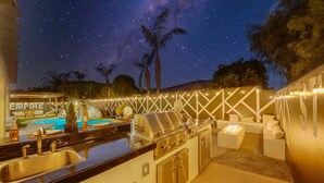 An outdoor kitchen area with a grill, sink, and bar seating, surrounded by string lights and palm trees under a starry night sky. - Empire State 7br, Golf, Waterslide, Gamerm (Indio)