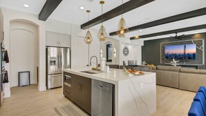 A modern kitchen with stainless steel appliances, a dishwasher, and a large island with a sink. There is a seating area with a sofa and a TV mounted on the wall. - Empire State 7br, Golf, Waterslide, Gamerm (Indio)